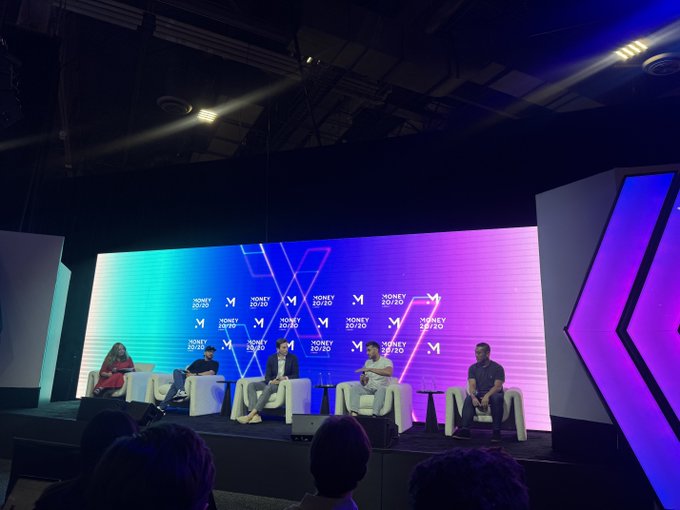 Five people sit in white chairs on a stage during a 'MONEY 20:20' panel discussion, with an illuminated blue and purple screen behind them.