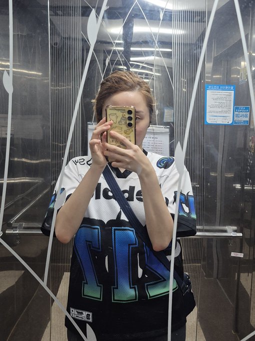 A mirror selfie of a person wearing a white and blue jersey with the number 14, holding a phone with a gold case in an elevator.