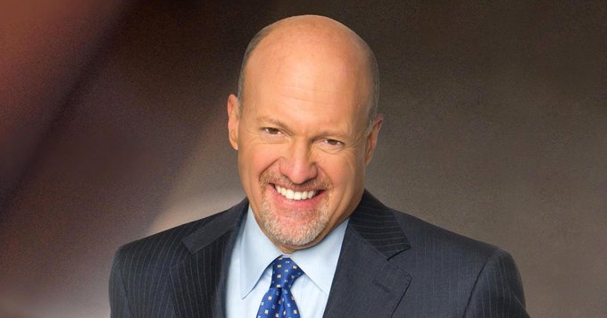 A professional headshot of a bald man with a gray goatee, smiling broadly, wearing a dark pinstripe suit, light blue shirt, and blue polka-dot tie against a brown background.