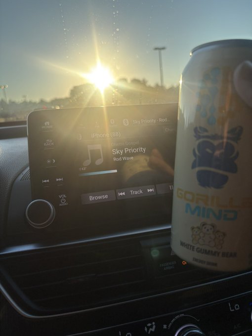 A photograph taken from the passenger seat of a car at sunset, showing the infotainment screen playing 'Sky Priority' by Rod Wave alongside a can of Gorilla Mind White Gummy Bear energy drink.