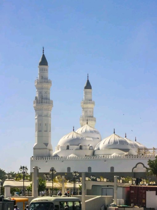 A white mosque with two tall minarets and multiple domes stands against a clear blue sky, with street lamps and vehicles visible at the base.