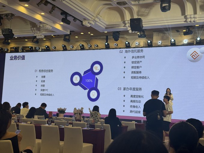 Conference or business presentation scene showing a large screen displaying Chinese text about business services (税务综合服务, 海外信托服务, 家办年度服务) with an audience seated in a banquet hall.