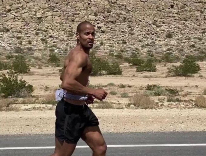 A shirtless, muscular man jogs along a road in a desert landscape, wearing black shorts and a white towel tucked into his waistband.