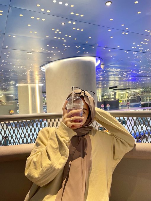 The image features a woman wearing glasses and a hijab standing next to a railing with her arm up. She has a drink in one hand, possibly a cup of coffee or tea, while holding a water bottle in the other hand. The background shows an indoor setting with a ceiling that is covered in lights, creating a bright atmosphere. There are also several people visible in the image, suggesting it might be a public space like a mall or a busy street.