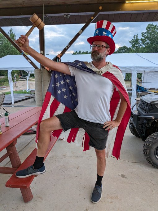 A man in an Uncle Sam costume, wearing a top hat and flag cape, posing playfully with a wooden gavel at an outdoor pavilion.
