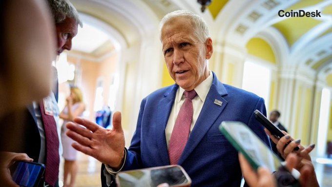 A man in a blue suit and pink tie speaks with reporters holding smartphones and recording devices in a government building corridor, with a "CoinDesk" watermark in the upper right. The image illustrates the article's focus on political negotiations surrounding the CLARITY Act, as lawmakers work to resolve ethics concerns before the White House decision deadline to push crypto legislation through before the recess.
