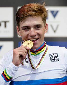 A cyclist wearing a white rainbow-striped world champion jersey smiles while biting a gold medal marked "UCI," with a "BMC Swiss" logo visible on his chest. The image relates to the post because it shows Remco Evenepoel, the rider on whom a $102,000 bet has been placed to overtake Pogačar's Tour de France lead.