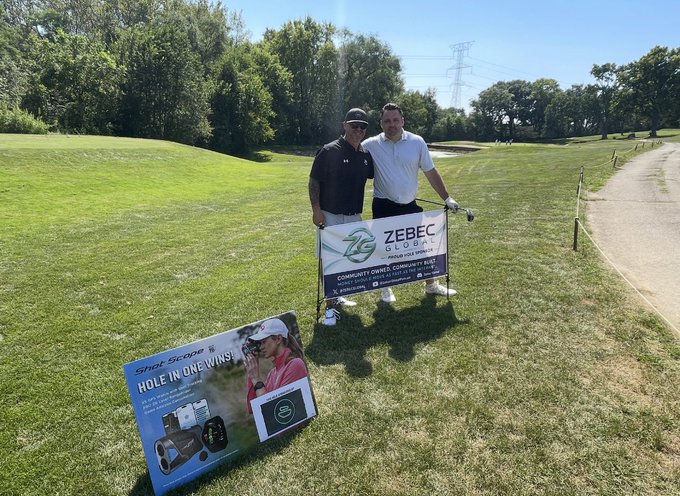 Two men posing on a golf course holding a 'ZEBEC GLOBAL' banner for a Community Owned Company, with a Shot Scope golf product display visible in the foreground.