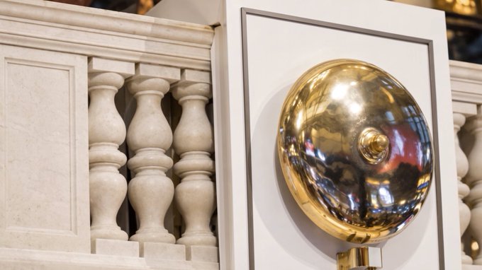 A polished golden dome or bell-shaped fixture mounted on a white framed panel, positioned next to a decorative white stone balustrade with classical balusters.