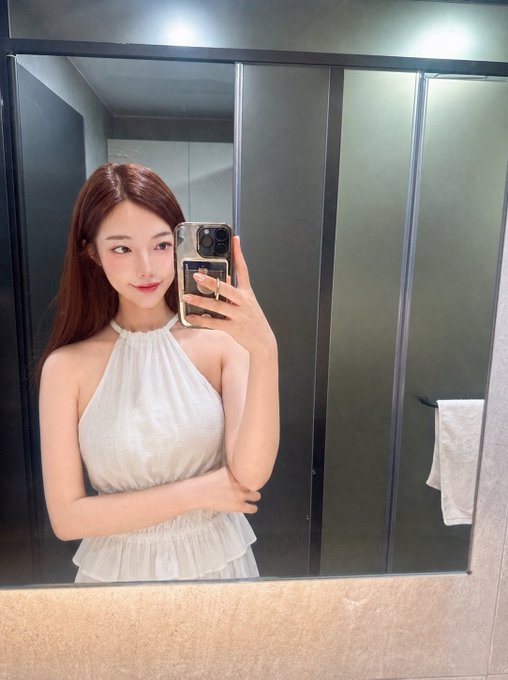 A mirror selfie of a young woman with long brown hair wearing a white halter top, holding a smartphone in a modern bathroom setting.