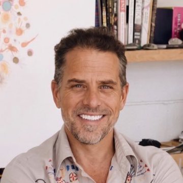 The image features a man with a beard wearing a shirt that has a floral pattern. He is smiling at the camera while sitting next to a bookshelf filled with various items such as books, a vase, and a clock. The background of the photo includes a wall adorned with colorful designs or paintings. The caption provided in the image seems unrelated to the man's appearance or the setting, but it does contain some text that is not directly related to the main subject matter.