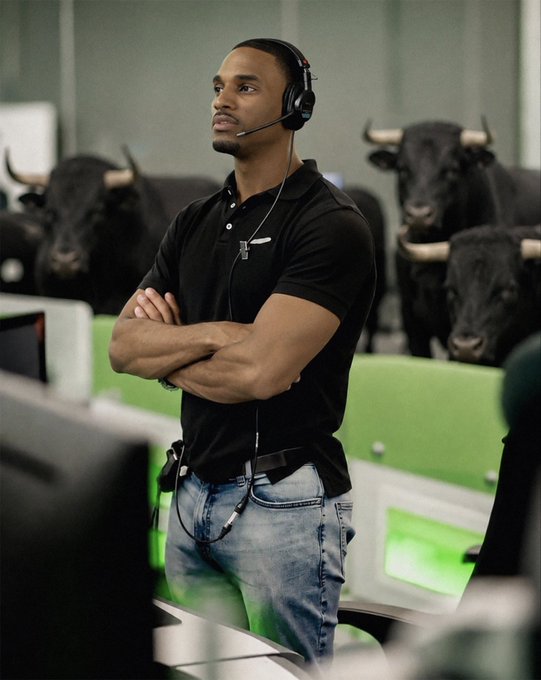 A man wearing a headset, black polo shirt, and jeans stands with crossed arms in a workspace, with several black bulls visible in the background.