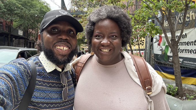 A selfie of a smiling couple on a city street, with the man wearing a black cap and patterned sweater, and the woman in a beige top with a brown backpack.