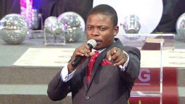 A man in a dark suit with a red tie and pocket square speaking passionately into a microphone while pointing forward, appearing to be a pastor or preacher at a church or religious event.
