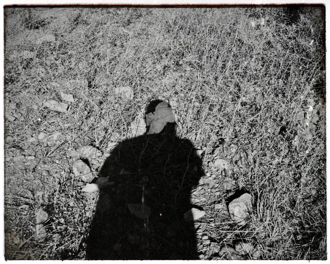 A black-and-white photograph with a vintage film border shows a person's dark silhouette/shadow cast onto rocky, brush-covered ground.