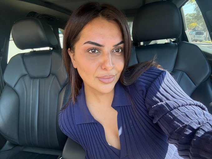 A woman sits in the driver's seat of a car wearing a navy blue ribbed sweater, taking a selfie with a neutral expression as sunlight streams through the windows. The image complements her post as a candid "good morning" update while she's on the go to finalize plans with the XRP community for 2027.