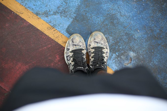 A first-person view looking down at a pair of white patterned sneakers standing on a blue and red surface with a yellow line.