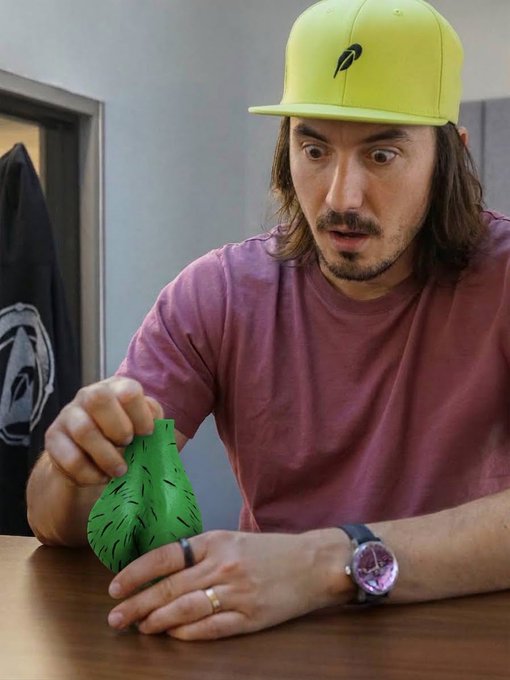 A man with long dark hair, wearing a neon yellow cap and a maroon t-shirt, looks with wide eyes at a small green creature inside a green paper bag on a wooden table.