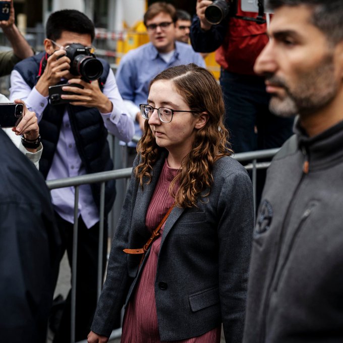 A young woman with glasses and a gray blazer walks past photographers and a man in a gray jacket, with metal barricades visible in the background. The image appears to relate to the post, likely depicting Caroline Ellison amid media coverage of the FTX-Anthropic share sale by the U.S. Marshals Service.