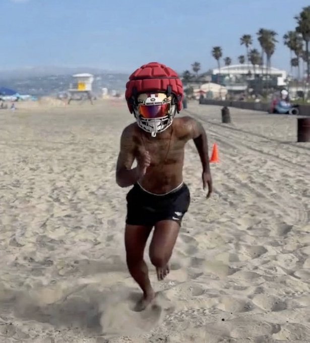 A shirtless man wearing a football helmet runs barefoot across a sandy beach, with palm trees and a lifeguard tower visible in the background. The image humorously visualizes the post's claim of "Romeo Doubs on the New England Patriots," depicting the Green Bay Packers receiver in an absurd beach training scenario.