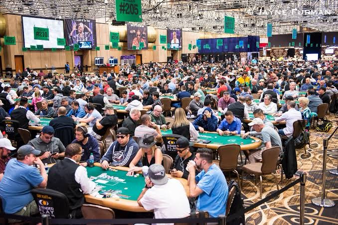 A crowded poker tournament hall with numerous players seated at green felt tables, featuring overhead screens displaying table numbers like '#205' and '#210', and signage reading 'HORSESHOE COUNCIL BLUFFS'.