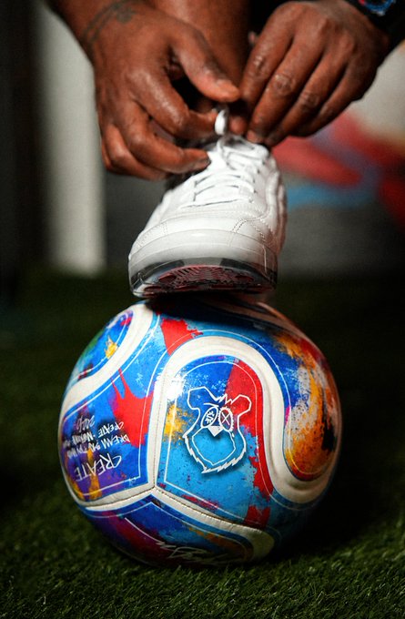 A close-up shot shows a person's hands tying the laces of a white sneaker, with the shoe resting on top of a vibrantly painted soccer ball on artificial grass.