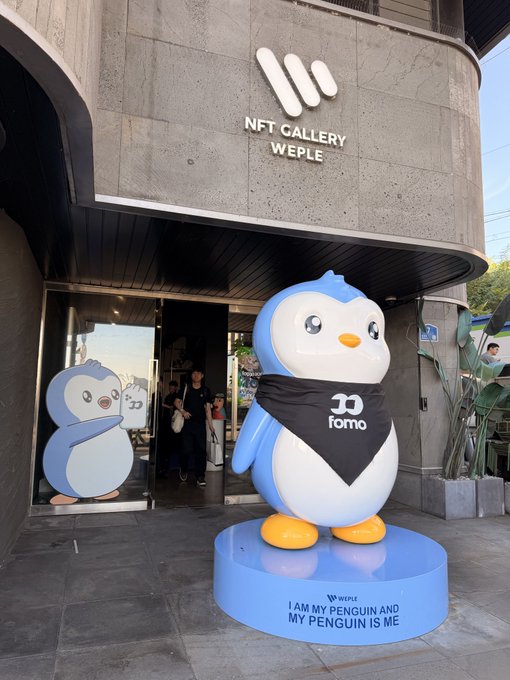 A large blue and white penguin statue wearing a bandana that says 'fomo' outside the 'WEPLE NFT GALLERY', with the base reading 'I AM MY PENGUIN AND MY PENGUIN IS ME'.