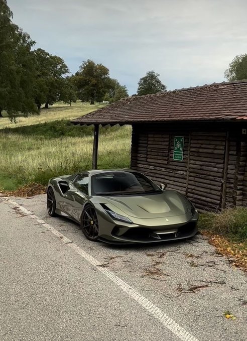 An olive green Ferrari sports car parked next to a rustic wooden cabin with a tile roof, surrounded by countryside scenery.