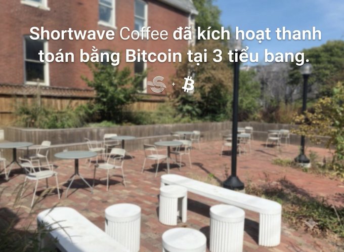An outdoor patio area with brick paving, tables, chairs, and modern white concrete benches, overlaid with Vietnamese text stating 'Shortwave Coffee đã kích hoạt thanh toán bằng Bitcoin tại 3 tiểu bang' (Shortwave Coffee has activated Bitcoin payments in 3 states), with Strike and Bitcoin logos.
