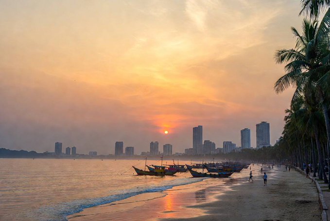 The image shows a beautiful sunset over a beach with several boats anchored near the shoreline, including small fishing boats. The sky has a warm orange hue as the sun sets behind the horizon. There are also people walking along the beach, enjoying the serene atmosphere and the view of the boats.