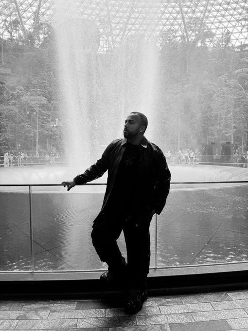 Black and white photo of a bearded man in dark clothing posing in front of an indoor waterfall feature, likely at Jewel Changi Airport.