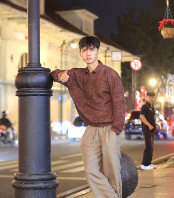 A young man in a brown sweater and beige pants leaning against a black lamppost on a city street at night.
