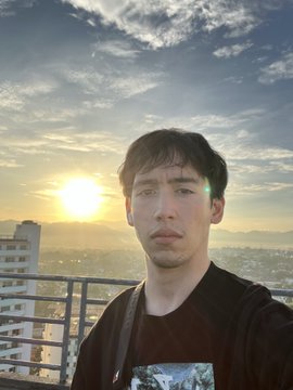 A selfie of a man with dark hair wearing a maroon shirt, taken outdoors with a sunset, sky, and distant cityscape visible in the background.