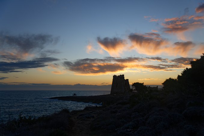 A scenic sunset over the ocean features a silhouetted stone tower on the rocky coastline against a sky with orange and blue clouds.