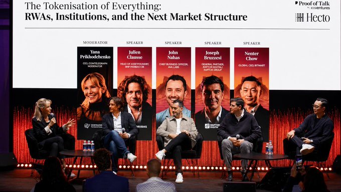 The image shows a group of people sitting around a table, with some standing behind them as they engage in conversation. The scene appears to be taking place at an event or conference where speakers are discussing various topics. There are several banners hanging on the wall behind the attendees, and one banner is specifically for the post of talk. The image also contains text that reads "The tokenisation of everything: RAVs, institutions, and the next market structure", which suggests a connection to the event or conference taking place in the image.