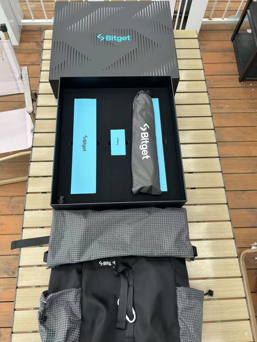 Bitget promotional package unboxed on a wooden deck, featuring a black box with teal 'Bitget' branding containing accessories and an umbrella, with a Bitget-branded apron displayed in front.
