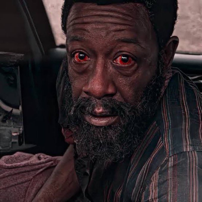 A close-up portrait of a bearded man with glowing red eyes and a worn, weathered expression, sitting inside what appears to be a vehicle.