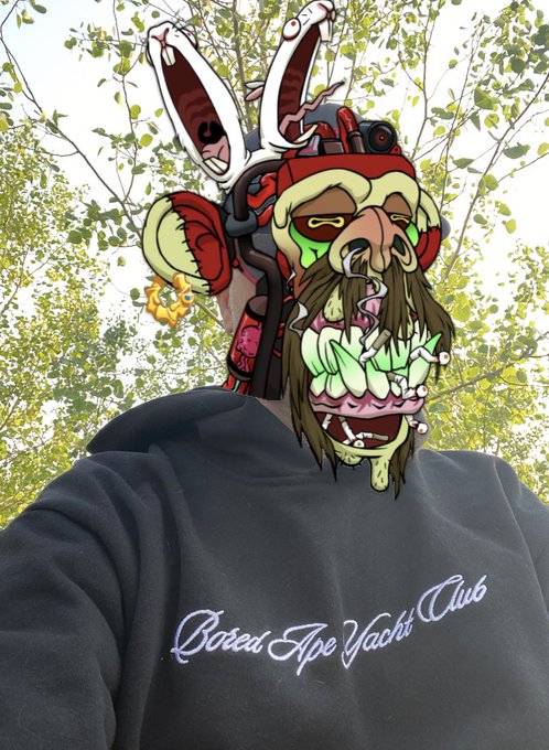 A person wearing a 'Bored Ape Yacht Club' hoodie stands outdoors under trees, with a grotesque cartoon zombie-ape face edited over their head featuring bunny ears.