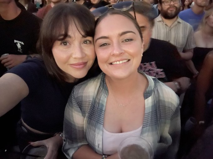 Two women smile closely together in a selfie taken at a crowded event, with one wearing a plaid shirt and the other a black top.