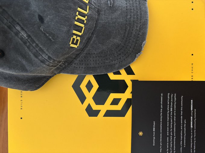 A dark, worn baseball cap with 'BUILT' embroidered on it rests on a yellow folder marked 'BUILT WEB' and 'PITCH DECK', alongside a black document about BNB Chain.