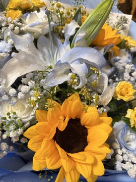 A close-up photograph of a colorful floral arrangement featuring a bright yellow sunflower, white lilies, baby's breath, and yellow roses.