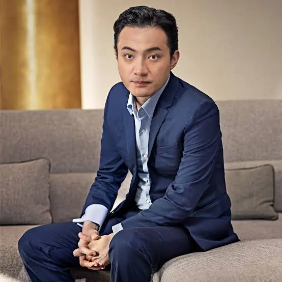 A man in a blue suit sits on a gray couch, hands clasped together, in a formal portrait-style photograph. The image appears to depict Justin Sun, the subject of the accompanying post about his procedural victory against World Liberty Financial.