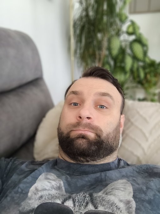 The image shows a man wearing a black shirt with cat ears sitting on a couch. The man has a beard, mustache, and glasses. He appears to be posing for a selfie or taking part in an online conversation about cats.