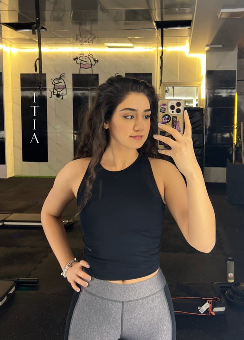 A woman with long dark hair takes a mirror selfie in a gym, wearing a black sleeveless crop top and gray leggings while holding a phone with a purple case. The "TITIA" sign and kettlebell graffiti art on the gym wall are visible in the background, fitting the post's motivational "UPtober" theme of fresh fitness energy.