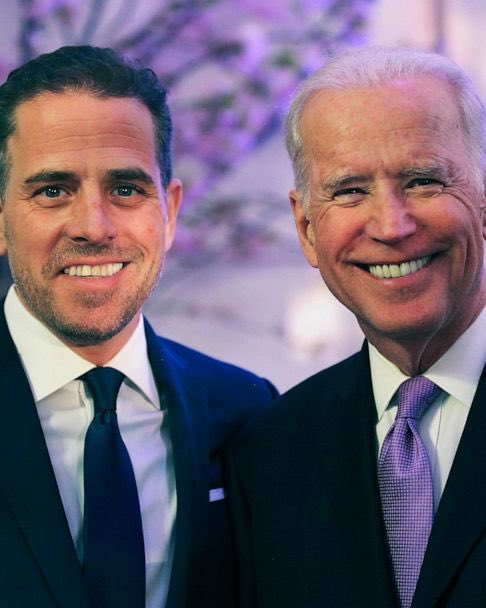 Two men in dark suits and ties pose for a smiling portrait, with one wearing a purple patterned tie and the other a black tie.