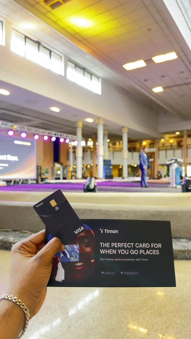 A hand holds a black Timon Visa bank card and a promotional pamphlet reading "THE PERFECT CARD FOR WHEN YOU GO PLACES" in a large indoor venue or conference hall.