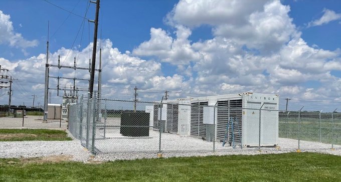 An outdoor industrial installation showing two large white Citronix brand air conditioning or cooling units secured inside a chain-link fence enclosure, set against a backdrop of green fields and a cloudy sky.