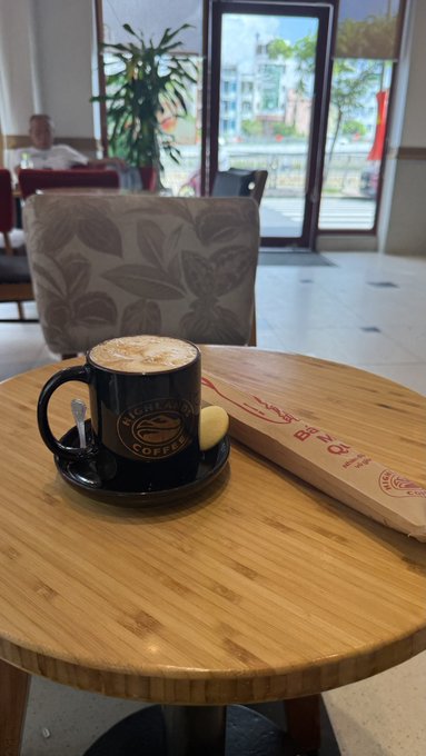 A black mug of cappuccino on a wooden table inside "Highlands Coffee," accompanied by a small biscuit and a brown paper bag with red Asian characters printed on it.