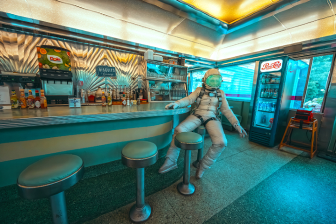 A digitally composited image depicts an astronaut sitting on a stool at a retro diner counter, with a Coca-Cola refrigerator and barista area visible in the background.