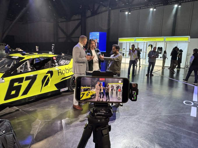 A professional interview is being filmed on a smartphone at an event, featuring a man in a gray suit speaking with two others in front of a yellow NASCAR-style race car displaying "67" and "Robinhood" branding. This appears to be footage of Robinhood ($HOOD) executives discussing the company's recent announcement of adding $25 million worth of Bitcoin ($BTC) to its corporate balance sheet.